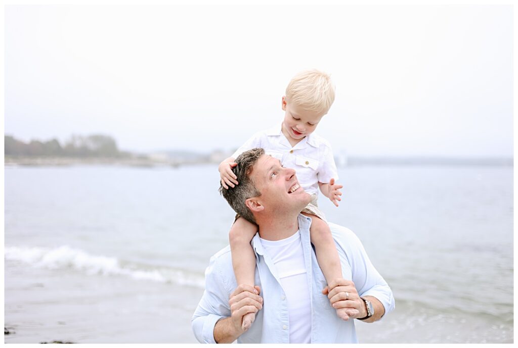 Father and toddler son wearing white and light blue standing by the ocean in NH during session with NH Family Beach Photographer