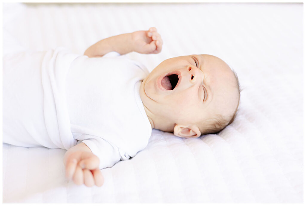 Newborn laying on a white bed in photography studio yawns and stretches. 