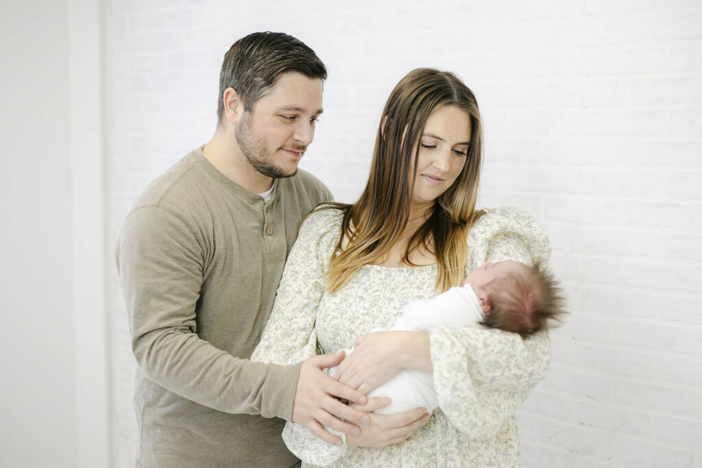 Mother and father holding their newborn baby swaddled in a white blanket during NH Photographer session. 